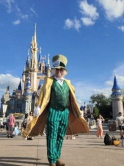 Greg Primrose as the Madd Hatter standing in front of an enchanted castle wearing a green suit, long gold coat, white gloves, and oversized checked top hat. He is smiling.